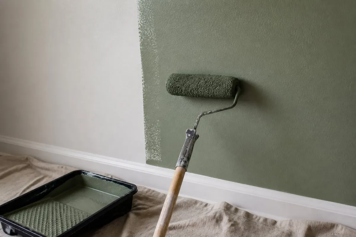A roller applying muted green paint to an interior wall above a protected floor.